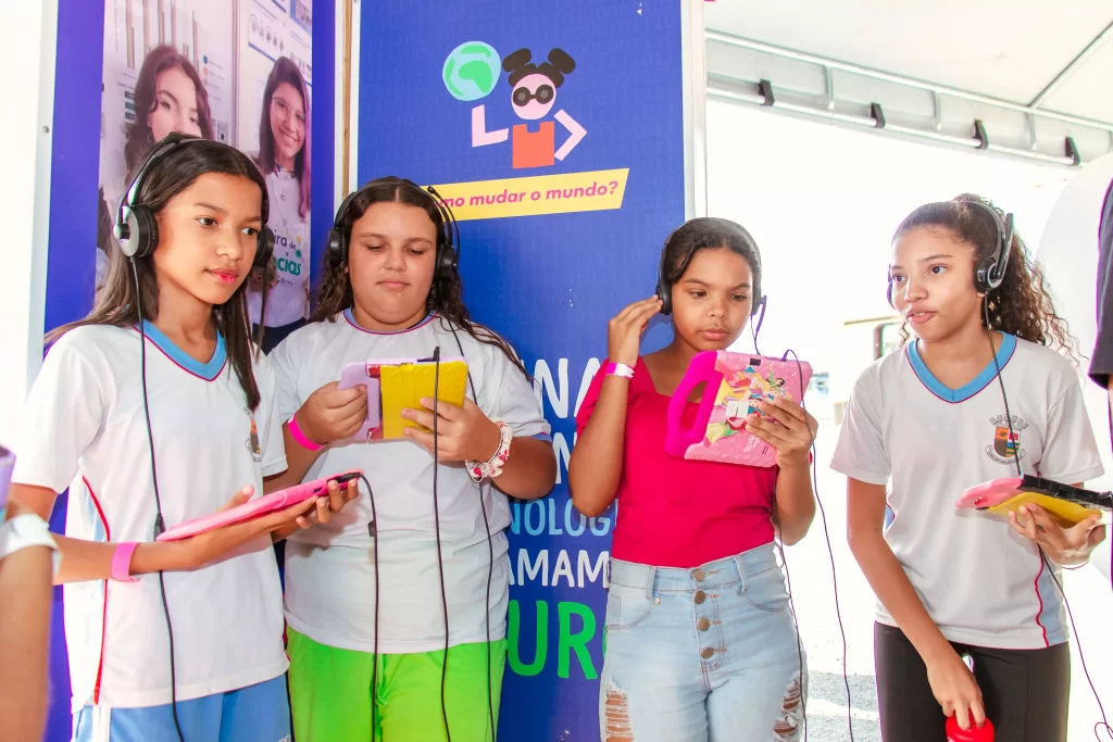 Um grupo de meninas em frente a um poster escutando histórias por um tablet